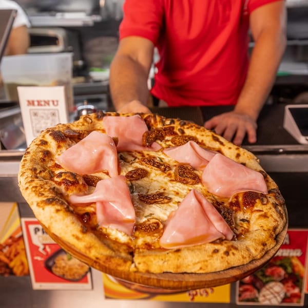 Chef in red shirt presenting a Pia's Not Just Pizza pizza with ham and figs.