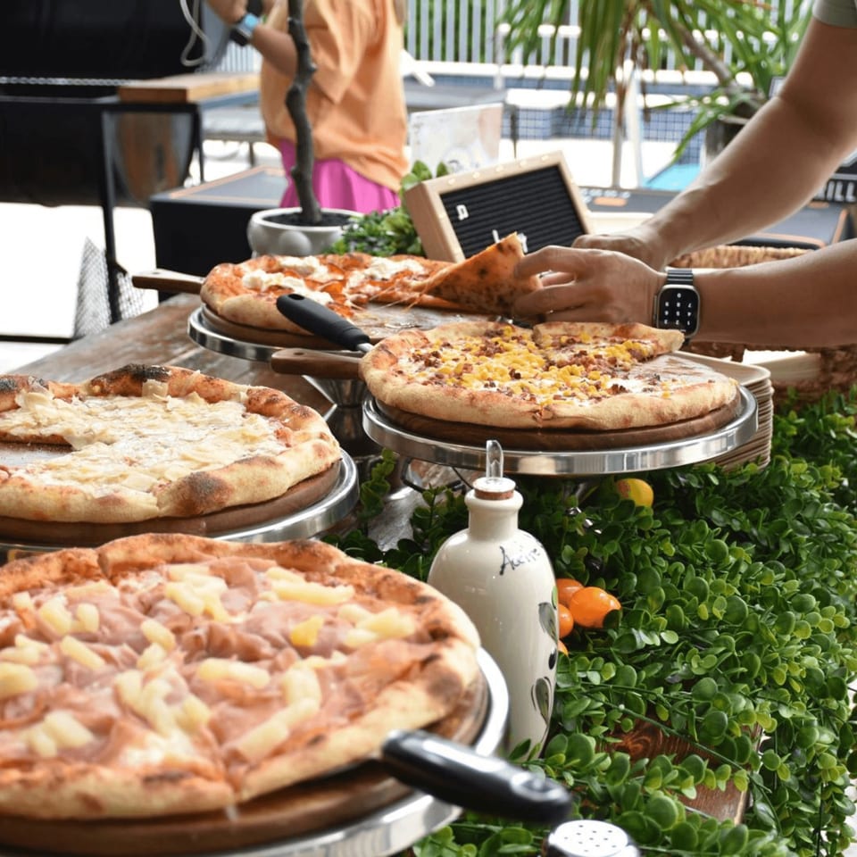 A person serving a slice of pizza from a buffet of various Pia's pizzas.