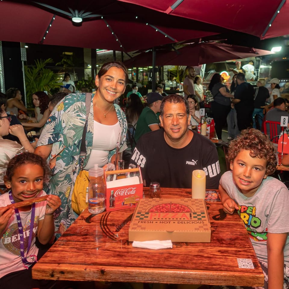 A family of four smiles at an outdoor table with a pizza box and drinks at Pia's Not Just Pizza.