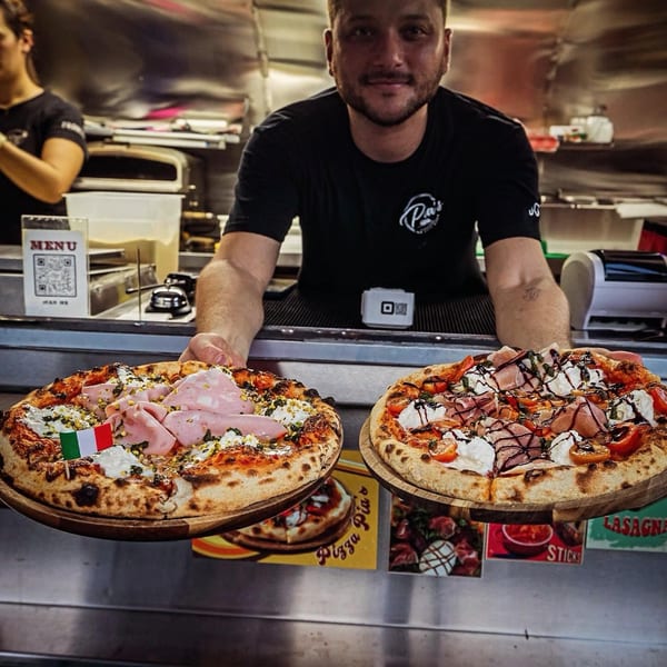 A chef from Pia's Not Just Pizza presenting two gourmet Italian pizzas with various toppings.