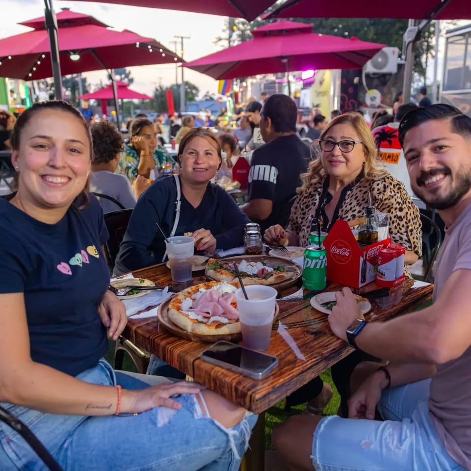 Four happy people enjoying pizzas and drinks at an outdoor table under red umbrellas at Pia's.