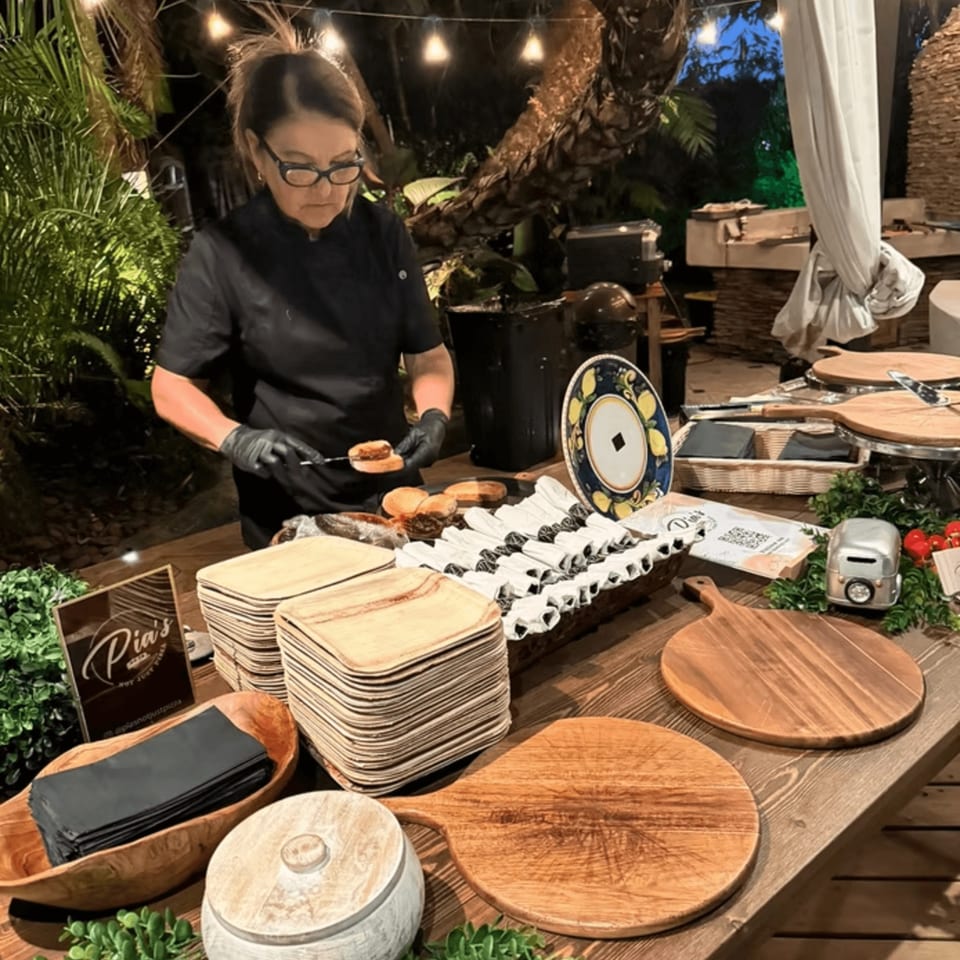 A chef in black uniform and gloves prepares food at an outdoor Pia's Not Just Pizza buffet table.