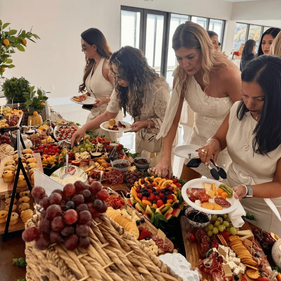 Women serving themselves from a large buffet table laden with fruits, cheeses, meats, and pastries.