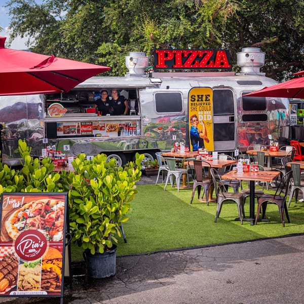 Pia's Not Just Pizza food truck with two employees, outdoor seating, and red umbrellas.