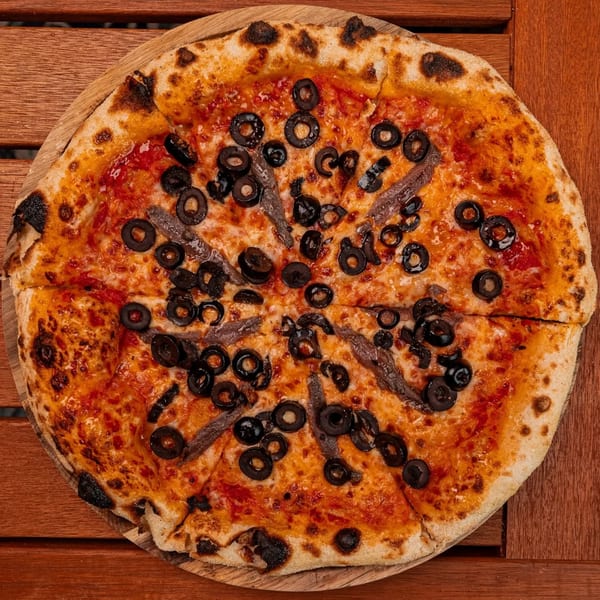 Pia's Not Just Pizza's sliced pizza with tomato sauce, cheese, black olives, and anchovies on a wooden board.