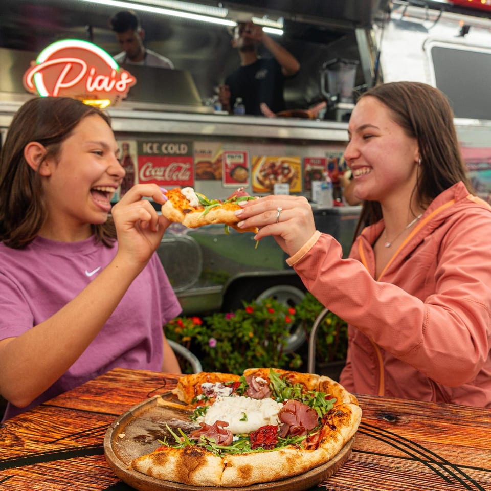 Two young women happily sharing a slice of Pia's pizza with burrata and prosciutto.
