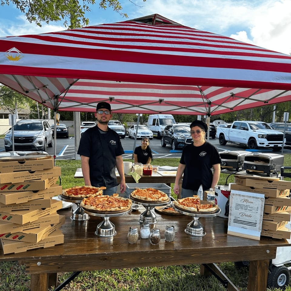 Pia's Not Just Pizza staff serving pizzas from a red and white striped tent at an outdoor event.